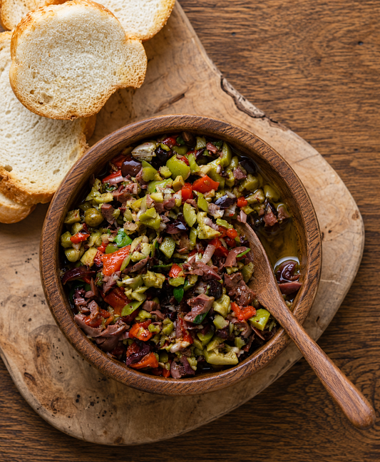 A wooden bowl filled Olive Bruschetta spread, set on a rustic wooden cutting board with slices of sourdough crostinis and a wooden spoon, on a dark wood table.